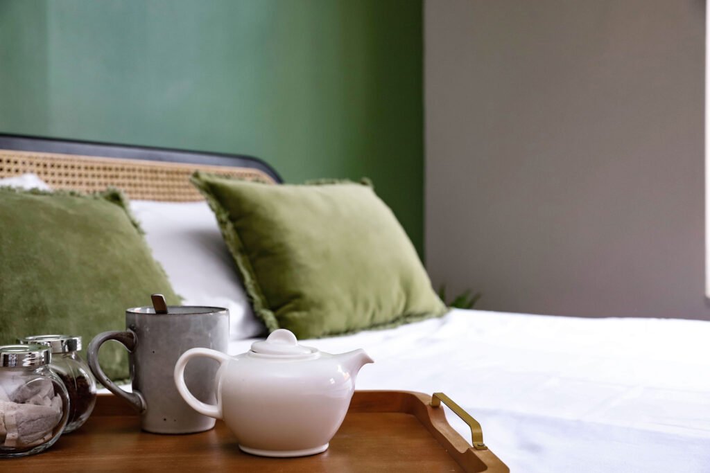 A tea pot accompanied by tea bags, sugar and mug on a wooden try on a bed.