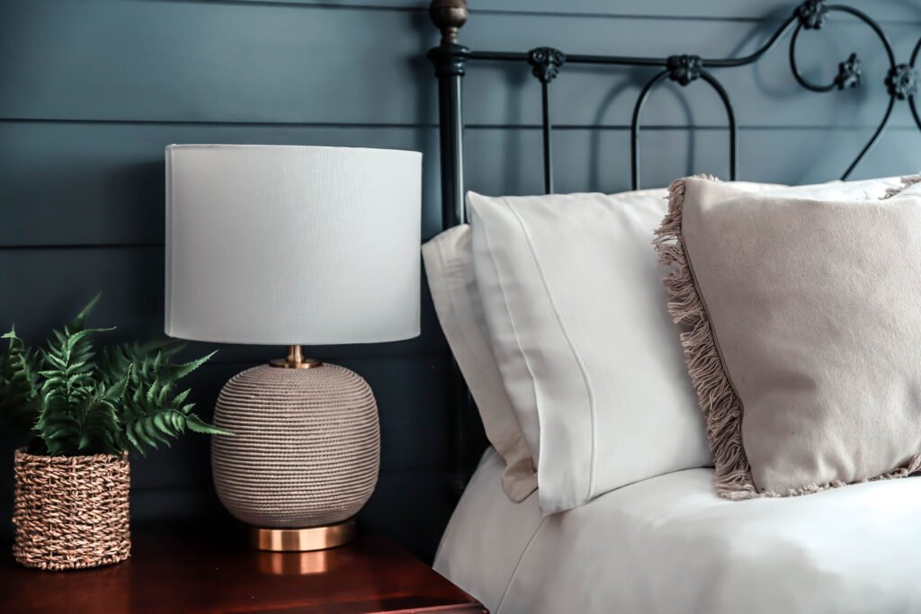 A bedside table with a plant and lamp rest next to a cosy bed.