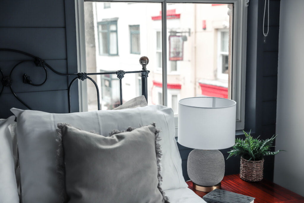 A grey and gold lamp resting on a bedside table next to a window.