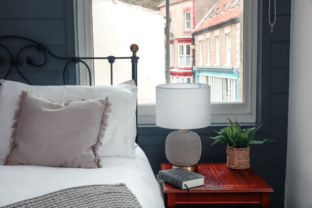 A grey and gold lamp on a bedside table accompanied by a reading book and green plant.